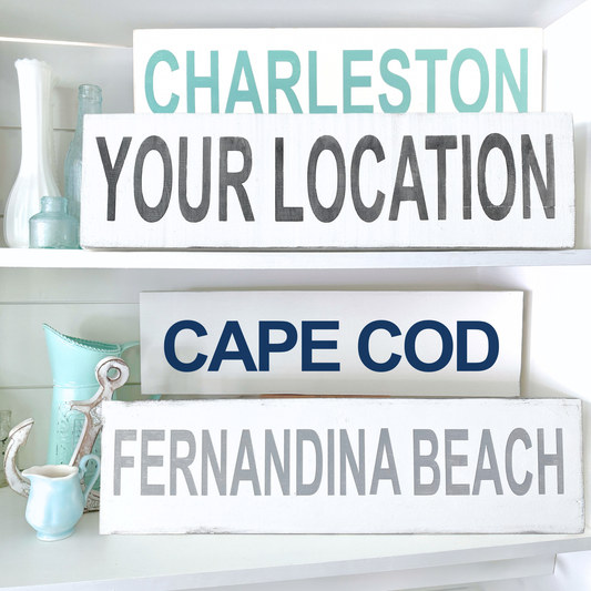 Four white rectangular wooden sign plaques displayed on white shelves, each with a different city or beach name: Charleston, Your Location, Cape Cod, and Fernandina Beach. The letters are in different colors: aqua, black, navy blue, and light gray.
