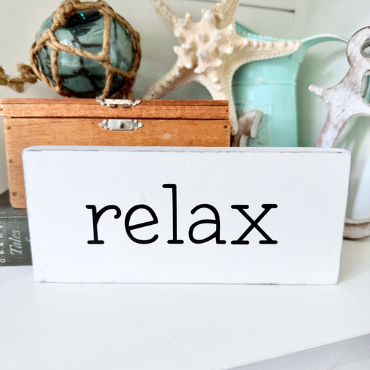 A white rectangular wooden sign with the word 'relax' in black lowercase letters, styled on a white surface with nautical decor in the background including a starfish, turquoise container, and rope ornament.