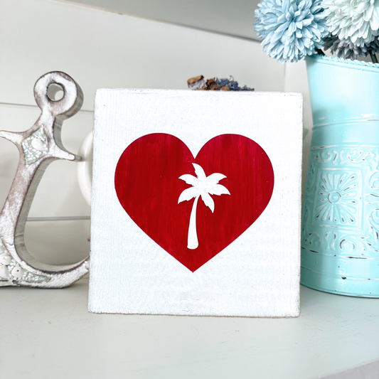 A square white wooden sign featuring a red heart with a white palm tree silhouette in the center, styled on a shelf next to a wood anchor and a light blue vase with flowers.