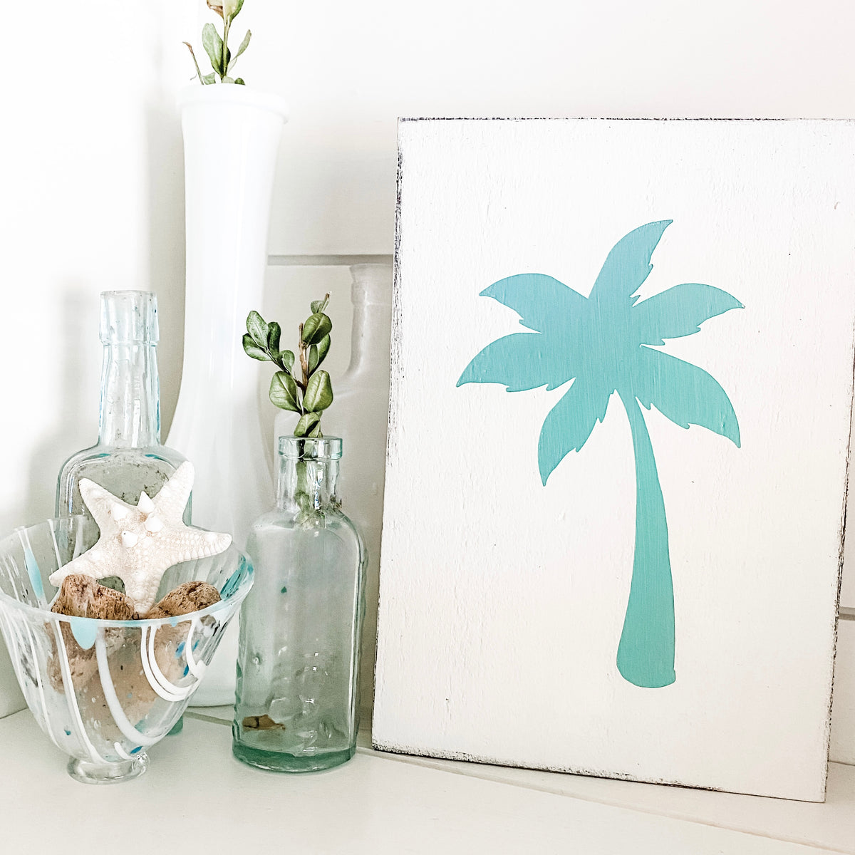 A white sign featuring a painted aqua palm tree silhouette, styled on a white surface next to glass bottles, a starfish, and a white vase