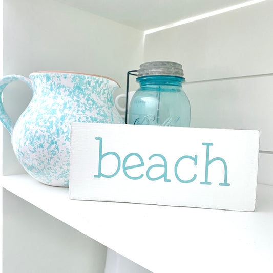A white rectangular sign with the word 'beach' in aqua, displayed on a white shelf next to a speckled blue-and-white pitcher and a blue glass mason jar.