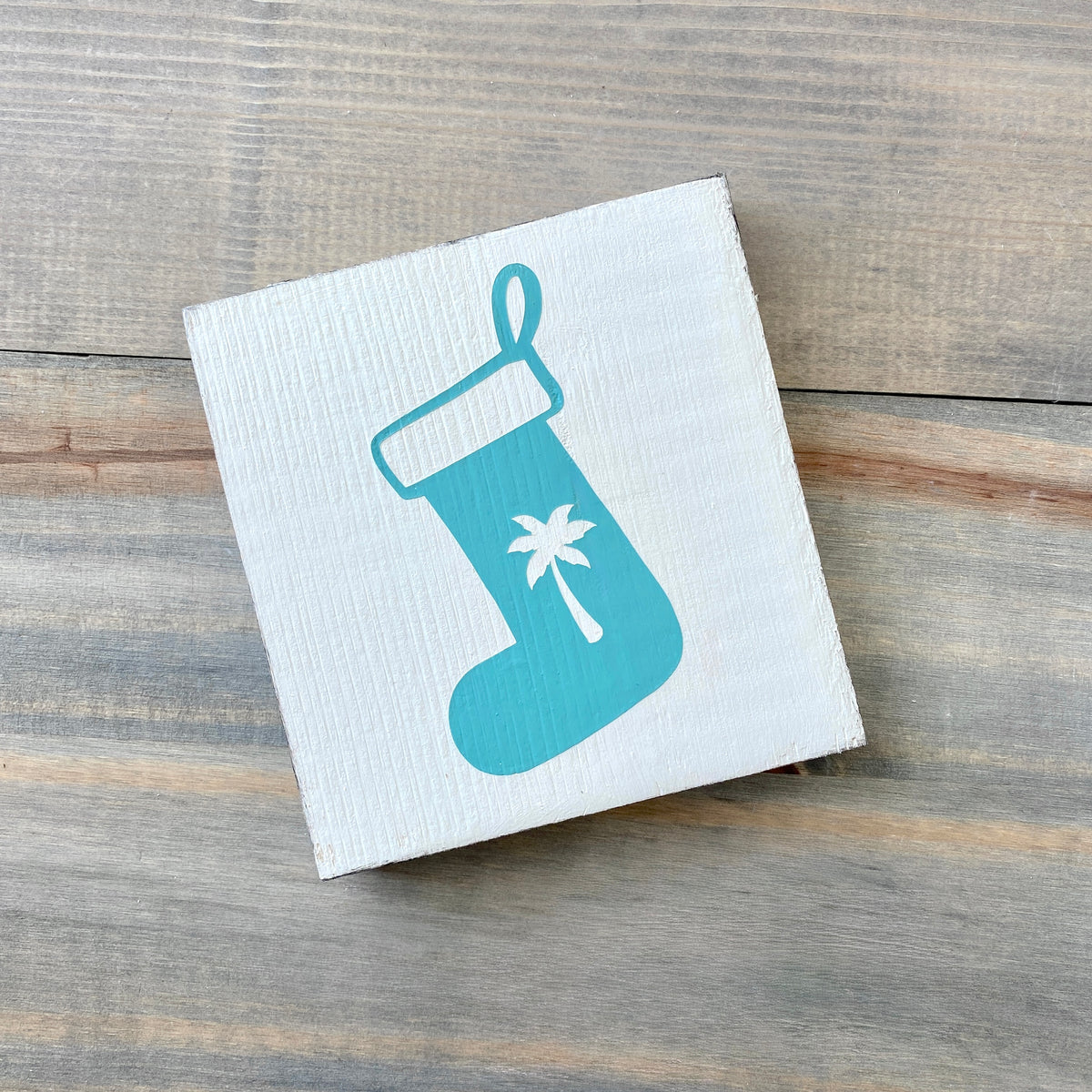 A square white wooden sign with an aqua Christmas stocking graphic featuring a white palm tree, displayed on a rustic gray wood plank surface.