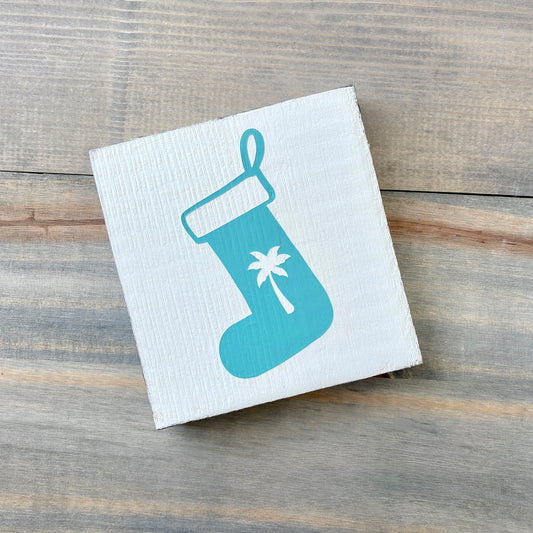 A square white wooden sign with an aqua Christmas stocking graphic featuring a white palm tree, displayed on a rustic gray wood plank surface.