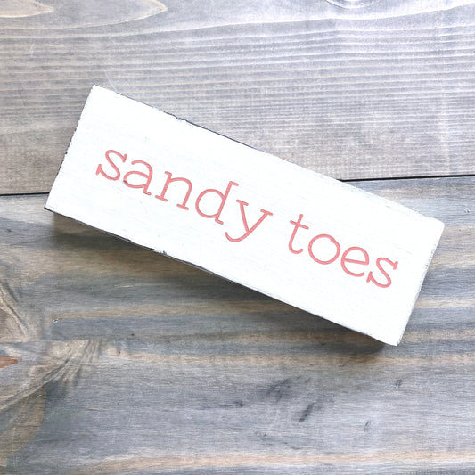 A rectangular white wooden sign with the words 'sandy toes' in a pale orange font, photographed on a rustic gray-brown wood plank surface.