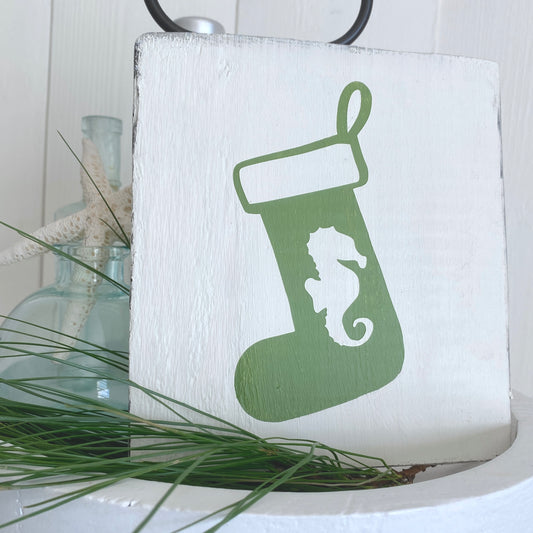 A square white wooden sign featuring a green Christmas stocking with a white seahorse silhouette, styled with green pine needles and a glass bottle in the background.