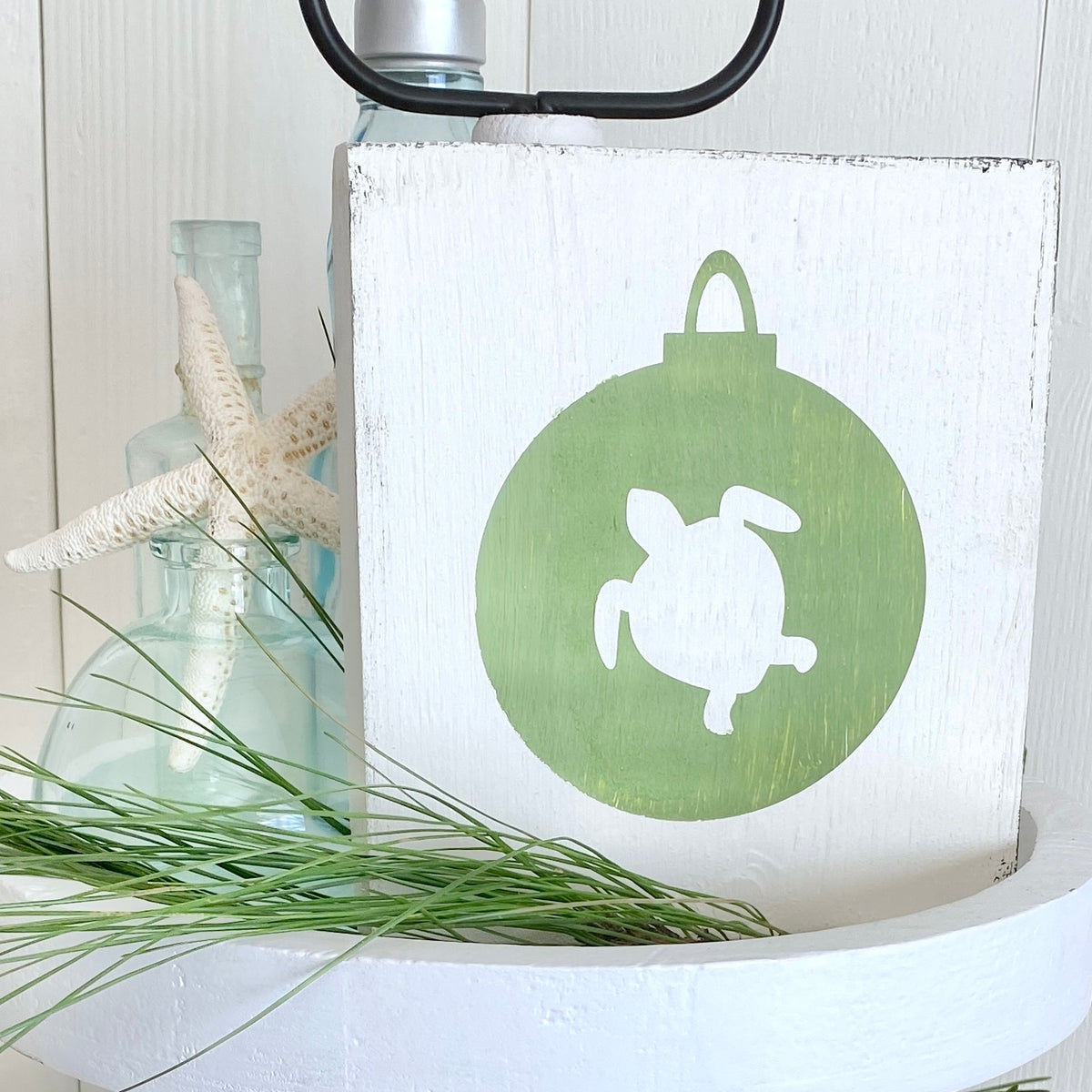 A rustic white wooden sign featuring a green circular ornament design with a white turtle silhouette. The sign is styled on a white pedestal with a starfish, a glass bottle, and green pine sprig against a white paneled background.