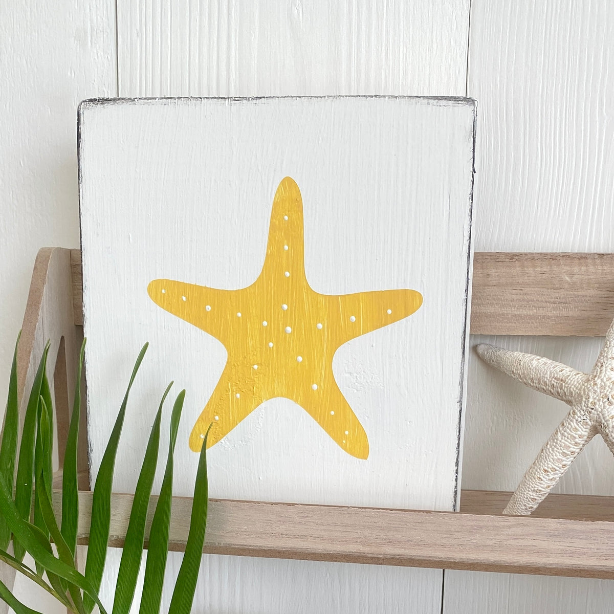 A square white wooden sign with a painted yellow starfish design, displayed on a wooden shelf with a green plant leaf in the foreground and a decorative starfish prop on the right.