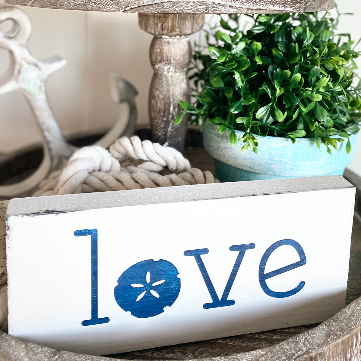 A white rectangular decorative sign with the word "love" in blue, where the letter "o" is replaced by a blue sand dollar motif. The sign is styled on a rustic surface with a rope coil, a potted green plant, and a blurred anchor in the background.