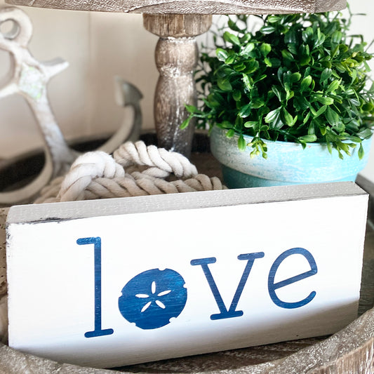 A white rectangular decorative sign with the word "love" in blue, where the letter "o" is replaced by a blue sand dollar motif. The sign is styled on a rustic surface with a rope coil, a potted green plant, and a blurred anchor in the background.