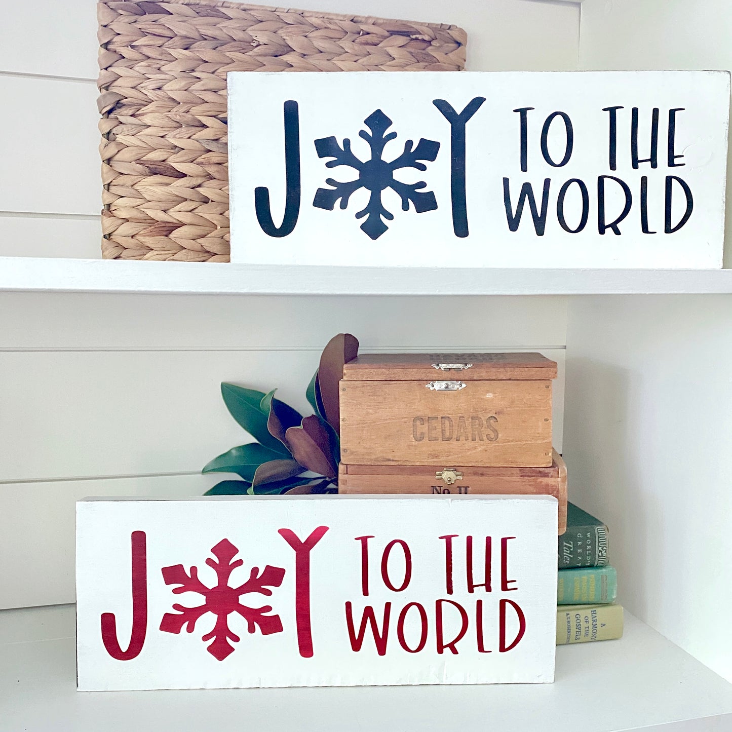 Two white decorative wall signs with the phrase “JOY TO THE WORLD” displayed on white shelves. The upper sign has dark black lettering and a snowflake, and the lower sign has dark red lettering and a snowflake.