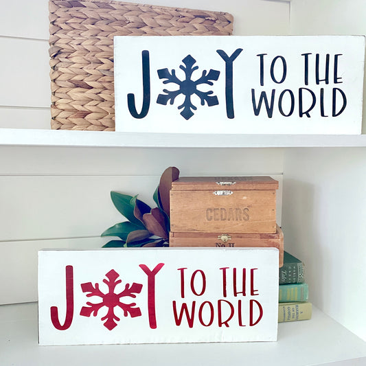 Two white decorative wall signs with the phrase “JOY TO THE WORLD” displayed on white shelves. The upper sign has dark black lettering and a snowflake, and the lower sign has dark red lettering and a snowflake.