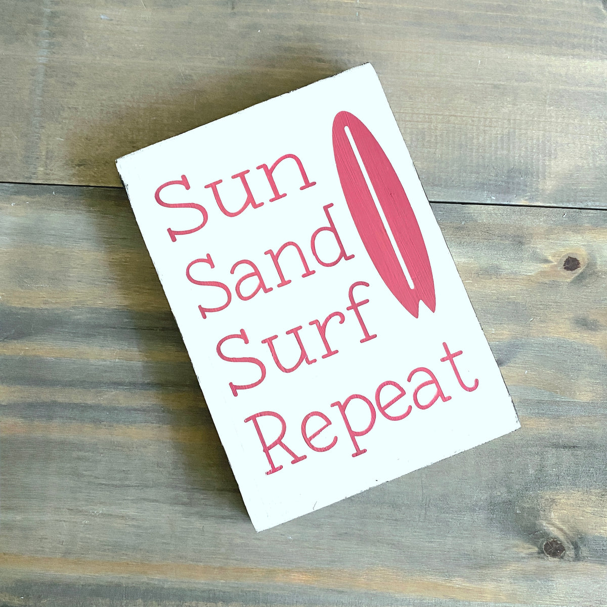 A white rectangular sign with the words 'Sun Sand Surf Repeat' in pink lettering and a pink surfboard graphic, placed on a rustic wooden surface.
