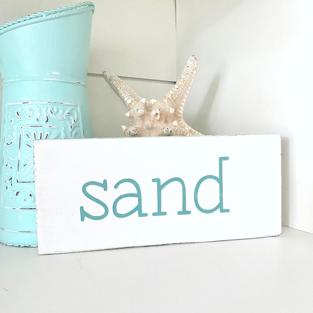 A white rectangular sign with the word "sand" in aqua lowercase letters, styled on a white surface with a turquoise metal container and a starfish behind it.
