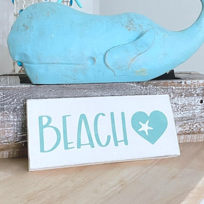 Blue whale figurine on a wooden surface with a 'BEACH' sign featuring a heart and starfish.