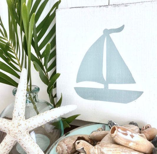 A white wooden sign featuring a sage green painted sailboat silhouette, styled with a white starfish in a glass vase, green leaves, and seashells in the foreground.