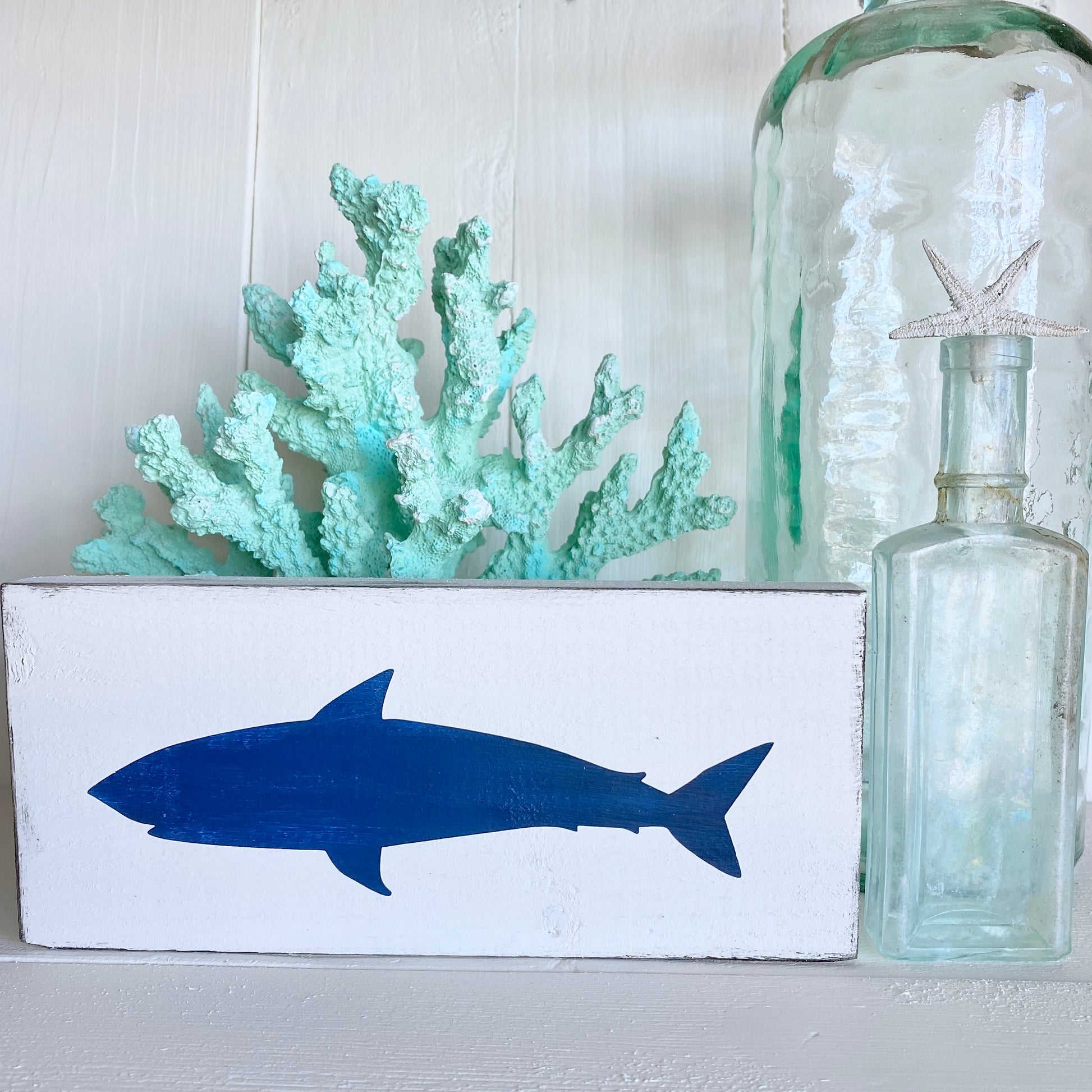 A white rectangular decorative sign featuring a navy blue shark silhouette, styled with a mint green coral piece and a large clear glass bottle on a white surface against a white paneled background.
