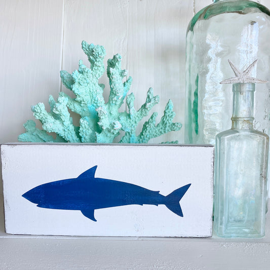 A white rectangular decorative sign featuring a navy blue shark silhouette, styled with a mint green coral piece and a large clear glass bottle on a white surface against a white paneled background.