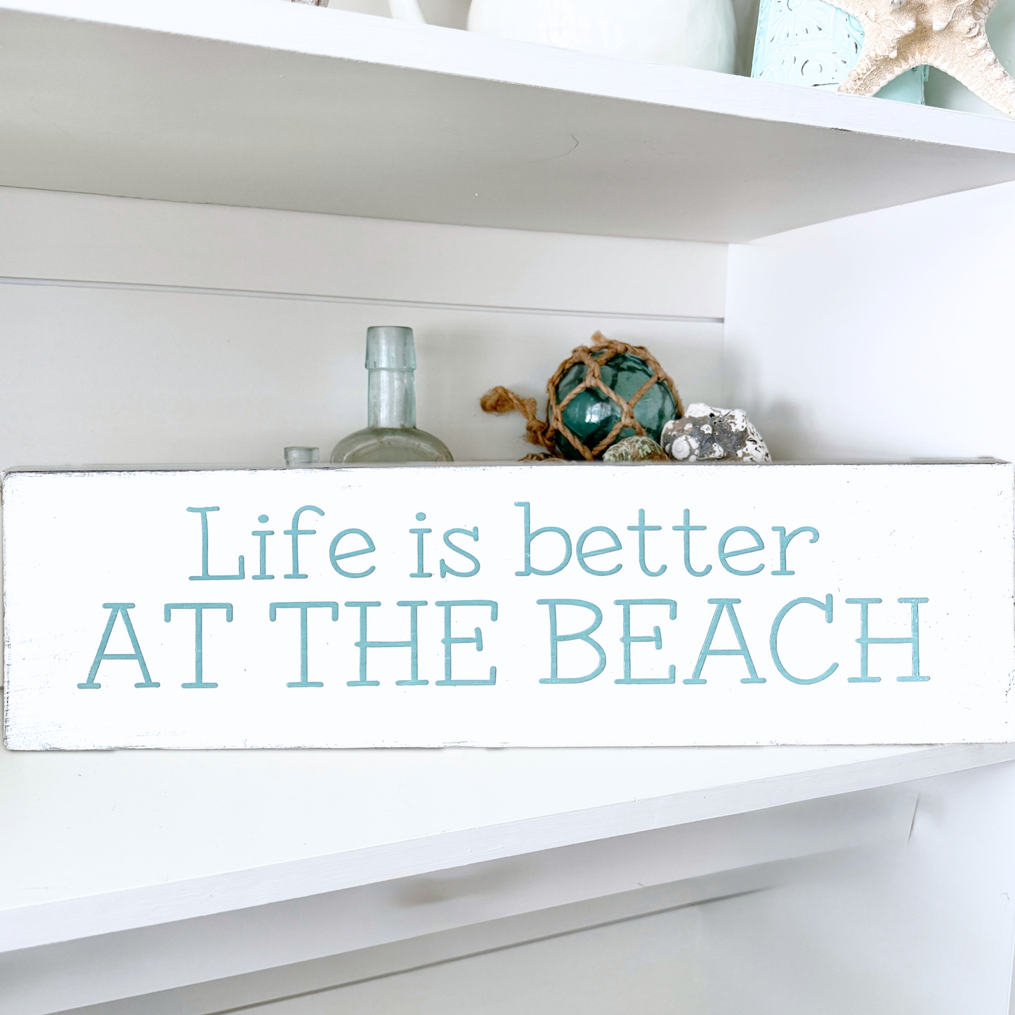 A white rectangular wooden sign with the phrase 'Life is better AT THE BEACH' in light blue lettering, displayed on a white shelf with decorative beach-themed items.