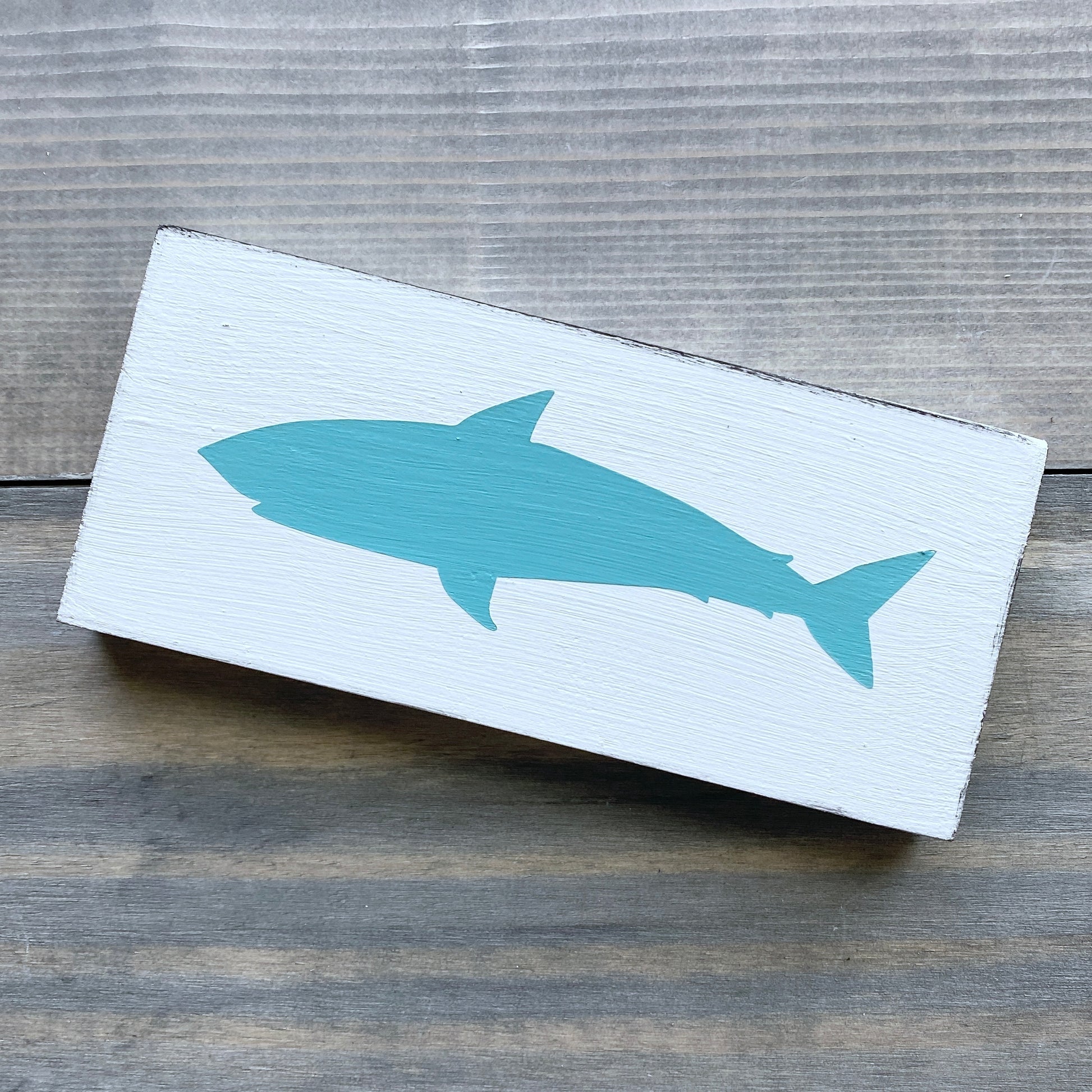 A rectangular white wooden sign featuring a painted aqua shark silhouette, displayed on a rustic gray wood plank surface.