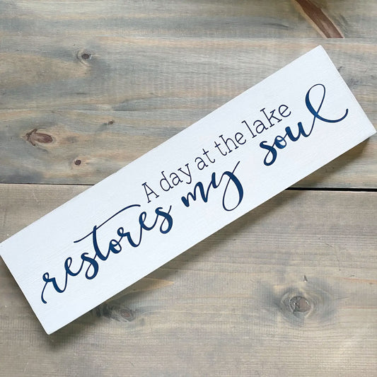 Wooden sign with text 'A day at the lake restores my soul' on a wooden surface