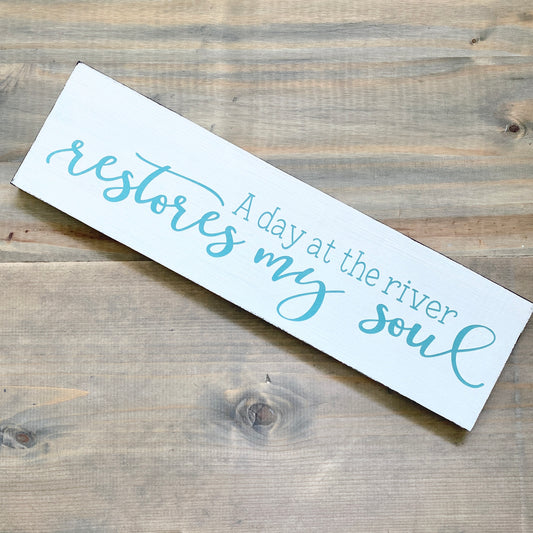 Wooden sign with text 'A day at the river restores my soul' on a wooden surface