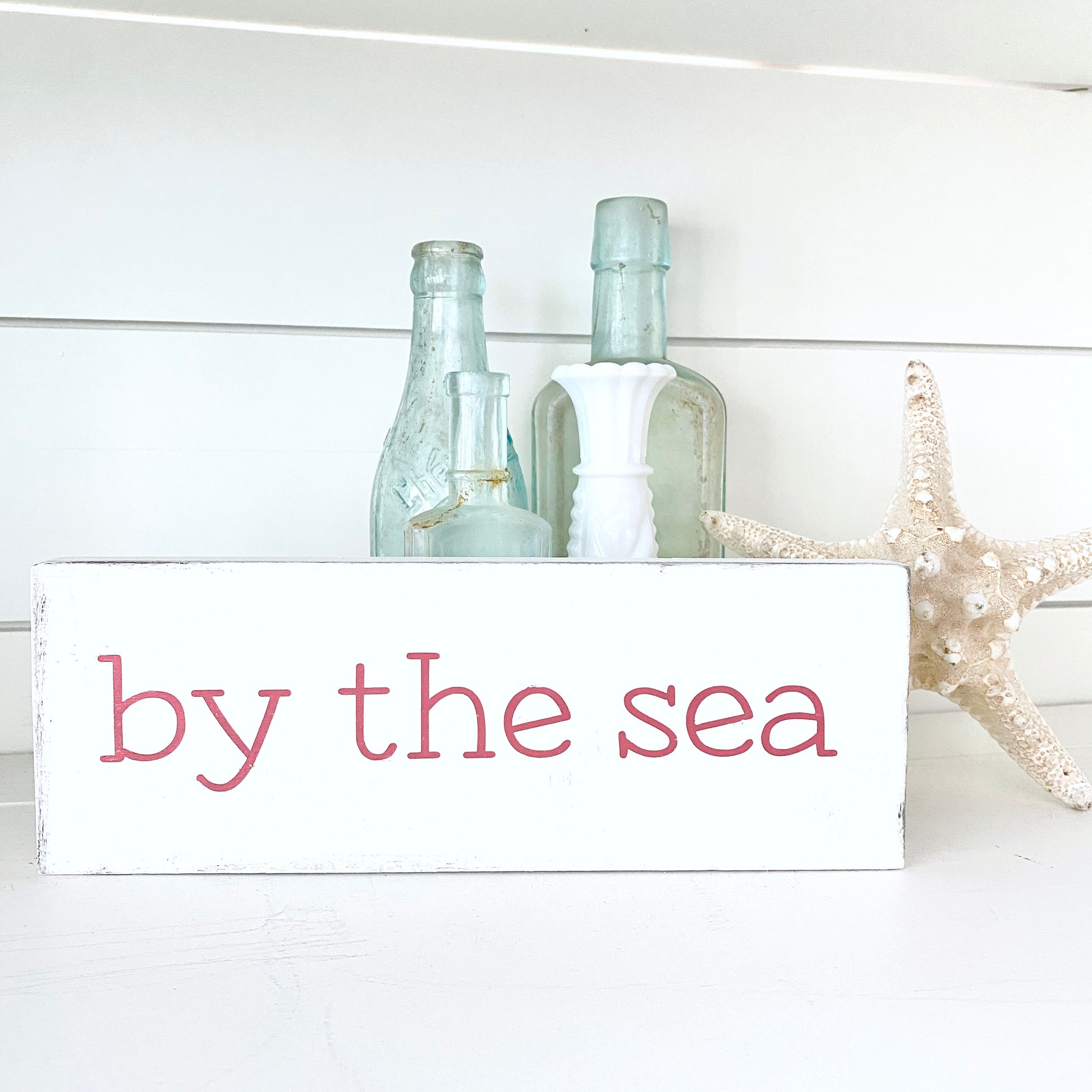 Decorative sign with 'by the sea' text, bottles, and starfish on a white background