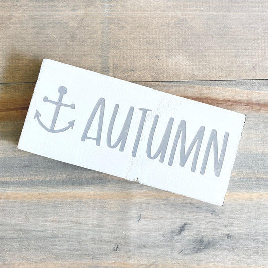 White sign with 'Autumn' and anchor design on a wooden surface