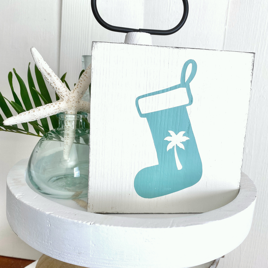 A square white wooden sign with an aqua Christmas stocking and white palm tree design, styled on a white tiered stand with a glass vase, starfish, and green leaves against a white paneled wall.