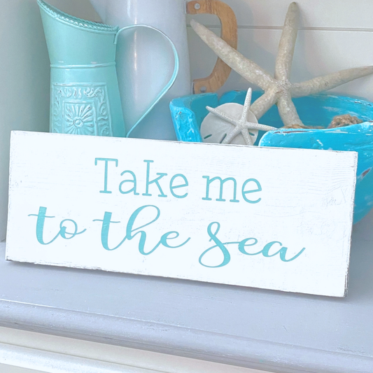 A white rectangular wooden sign with the phrase 'Take me to the sea' in aqua, styled on a shelf with turquoise decor, starfish, and a pitcher.
