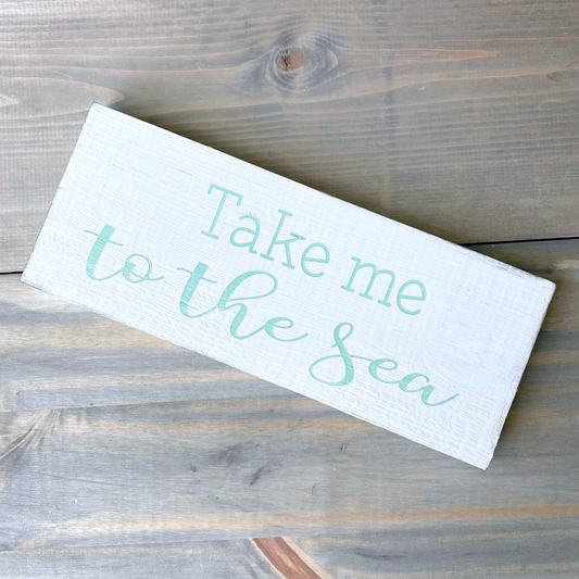 A rectangular white wooden sign with the phrase 'Take me to the sea' in mint script, photographed on a rustic gray-brown wood plank surface.