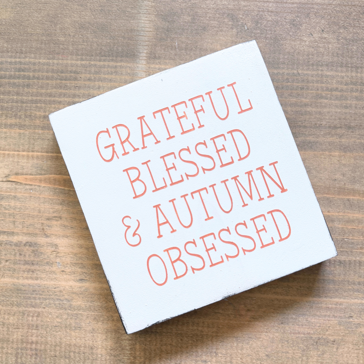 A square white decorative sign with the phrase 'GRATEFUL BLESSED & AUTUMN OBSESSED' in pale orange ettering, photographed on a brown wood surface.