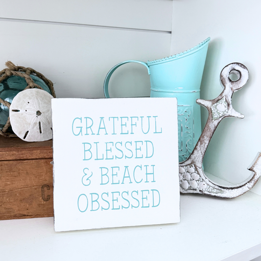 A white rectangular decorative sign with the phrase “GRATEFUL BLESSED & BEACH OBSESSED” in aqua lettering, styled on a white shelf with a turquoise pitcher, a distressed white anchor, and beach-themed decor.