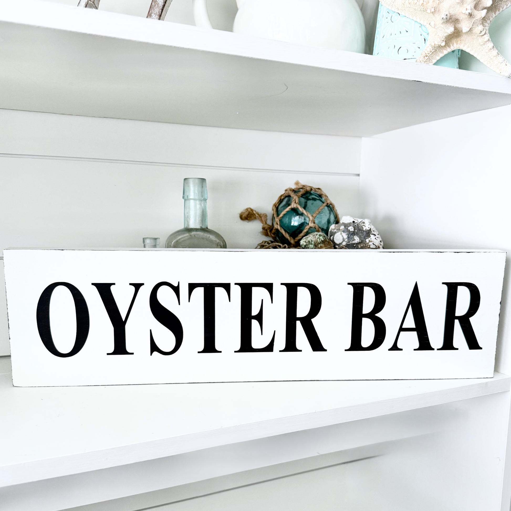 A white rectangular wooden sign with the words 'OYSTER BAR' in large black serif letters, displayed on a white shelf among decorative seashell and glass items.