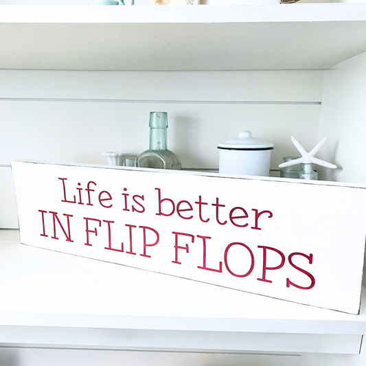 A white rectangular wooden sign with the phrase 'Life is better IN FLIP FLOPS' in red lettering, displayed on a white shelf alongside decorative home items.
