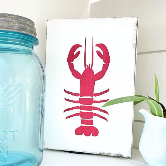 A rectangular white sign with a bright red 
lobster illustration, styled on a shelf next to a blue glass mason jar and a small potted plant.