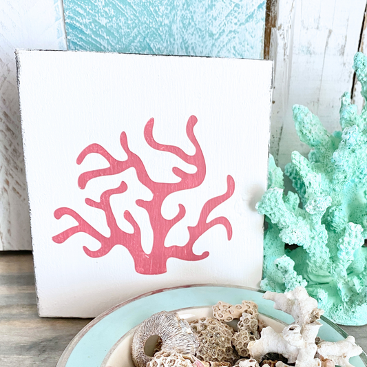 A square white sign featuring a coral branch design in pink, styled on a tabletop with coral pieces, a mint green decorative coral piece, and a distressed white wood background.
