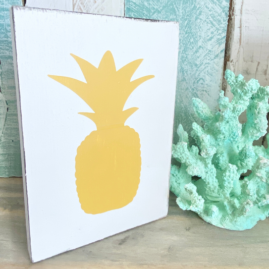 A white sign featuring a yellow pineapple silhouette, styled on a wooden surface next to a mint green coral decoration.