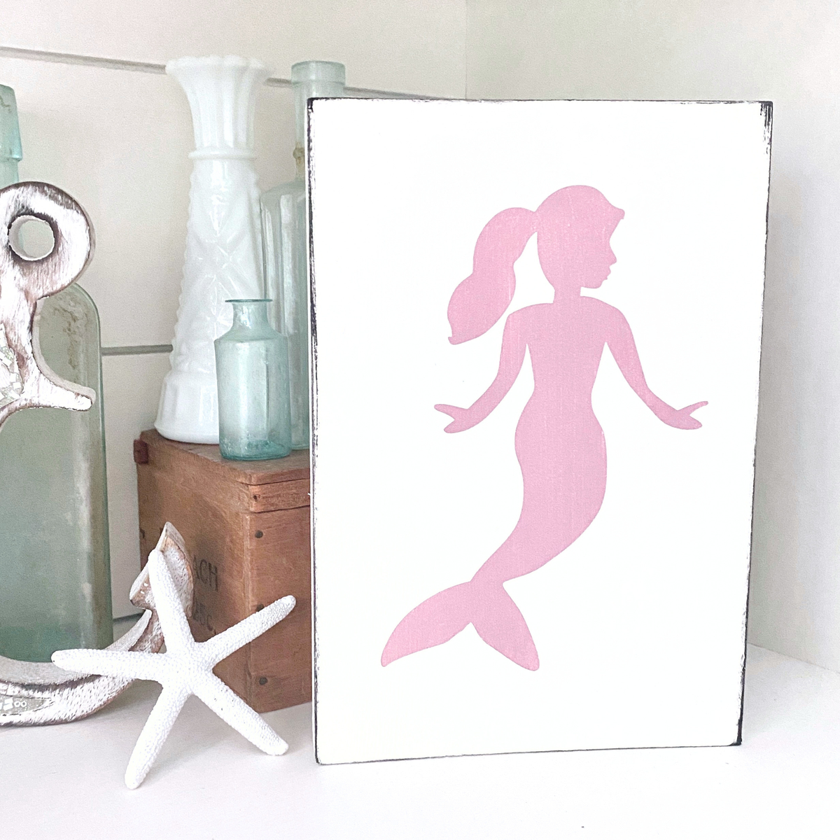 A rectangular white sign with a pink mermaid silhouette, displayed on a shelf next to decorative glass bottles, a starfish, and a white vase.