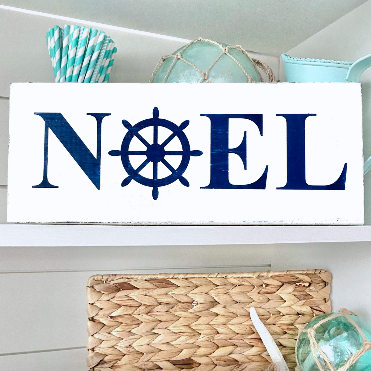 A white rectangular decorative sign with the word "NOEL" in dark navy blue, where the O is replaced by a ship's wheel motif. The sign is displayed on a white shelf with nautical-themed decor, including striped paper straws, a woven basket, and a light blue cup.