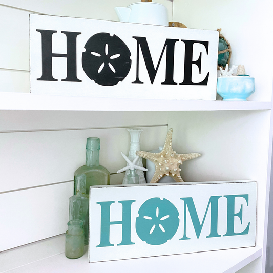Two white decorative wooden signs with the word “HOME” in black and aqua, styled on white shelves with seashell decor and glass bottles.
