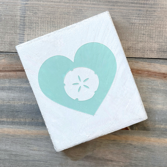 A square white sign wall art piece featuring a mint heart with a white sand dollar in the center, displayed on a rustic wooden surface.