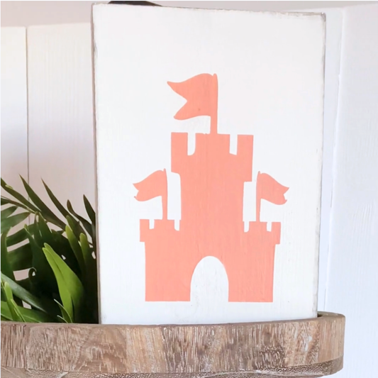 A rectangular white wooden sign featuring pale orange sandcastle silhouette with flags, displayed on a rustic wooden shelf next to a green plant against a white paneled wall.