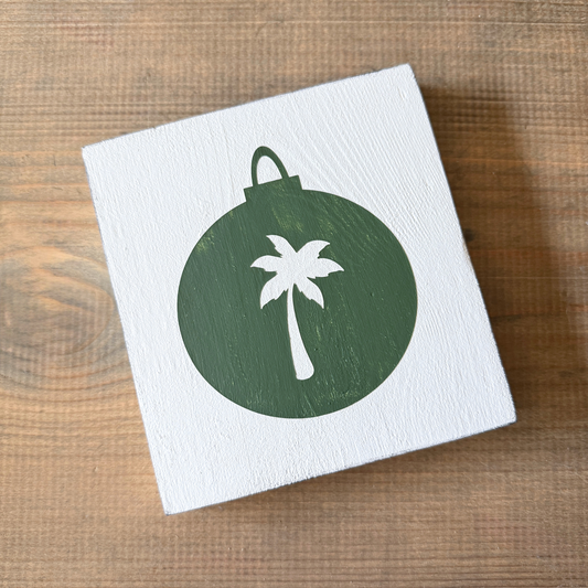 A square white wooden sign with a painted dark green ornament shape and a white palm tree design, photographed on a brown wood surface.