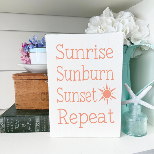 A white rectangular sign with the words 'Sunrise Sunburn Sunset Repeat' in pale orange lettering and a small sun icon, displayed on a shelf with decorative props including flowers, a wooden box, books, and a starfish.
