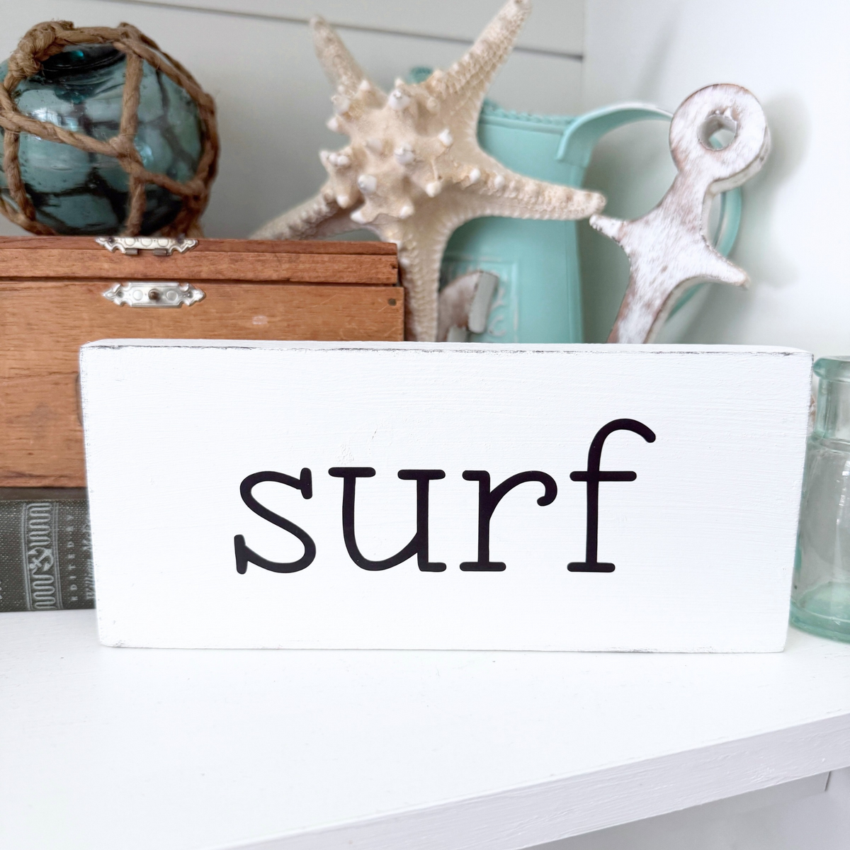 A white rectangular wooden sign with the word 'surf' in black lowercase, displayed on a white shelf with nautical decor including a starfish, turquoise pitcher, and wooden box in the background.