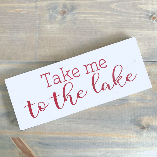 A white rectangular decorative sign with the phrase 'Take me to the lake' in red, photographed on a rustic wood plank surface.