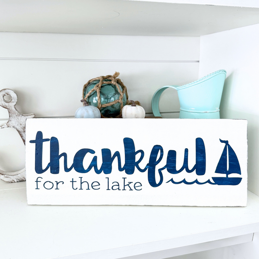 Thankful for the lake sign in white with navy lettering and sailboat and wave graphic. Placed on a white shelf with coastal and fall decor