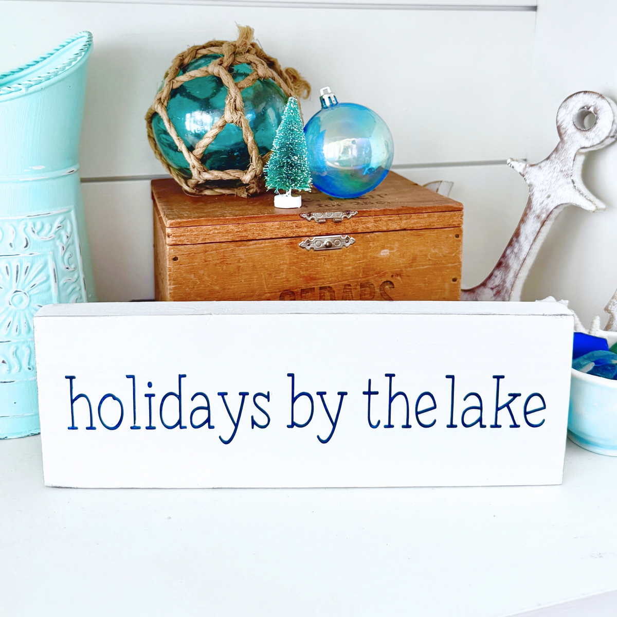 A white rectangular decorative sign with the phrase 'holidays by the lake' in navy, displayed on a tabletop in a nautical-themed setting with a wooden chest, blue ornaments, and an anchor.