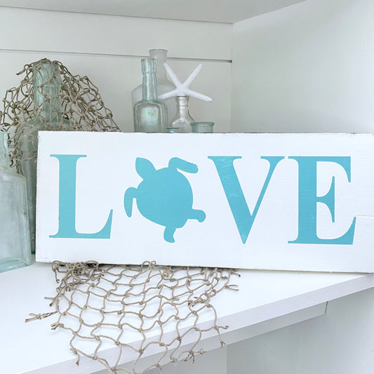 A white rectangular decorative sign with the word LOVE, where the O is replaced by aaqua sea turtle silhouette. The sign is styled on a white shelf with nautical decor, including glass bottles, a starfish, and netting.