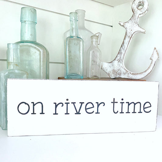 A white rectangular sign with the words 'on river time' in black lettering, styled on a shelf with vintage glass bottles and a distressed white anchor against a white shiplap background.